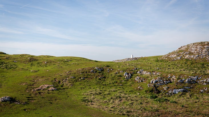 A grassy hilltop on Graig Fawr with scattered rocks. In the distance, a white trig point is visible.